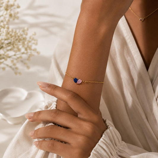 Dainty chain bracelet on a woman's wrist with a small heart formed by one blue and one pink birthstone color stone