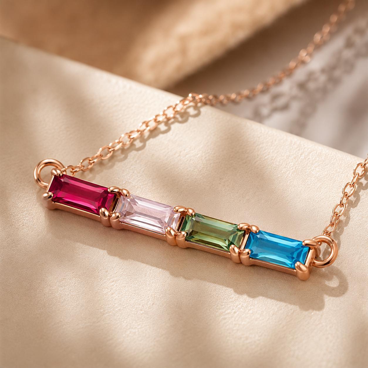 Close-up of a rose gold baguette birthstone necklace with four coloured baguette stones on cream fabric