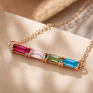 Close-up of a rose gold baguette birthstone necklace with four coloured baguette stones on cream fabric